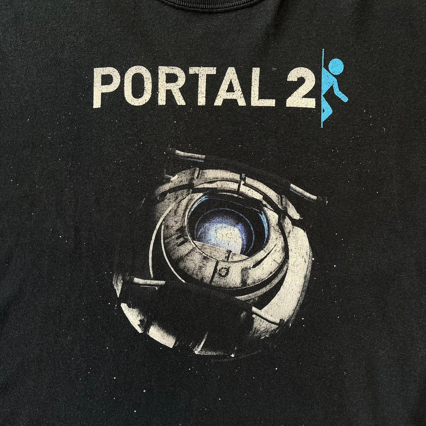 Portal 2: Wheatley in Space 2011 J!nx Tee features a black T-shirt made on a Gildan blank by J!NX in collaboration with Valve. The front has a graphic of Wheatley floating in space with a white and blue Portal 2 logo above him. Made from 100% cotton.
