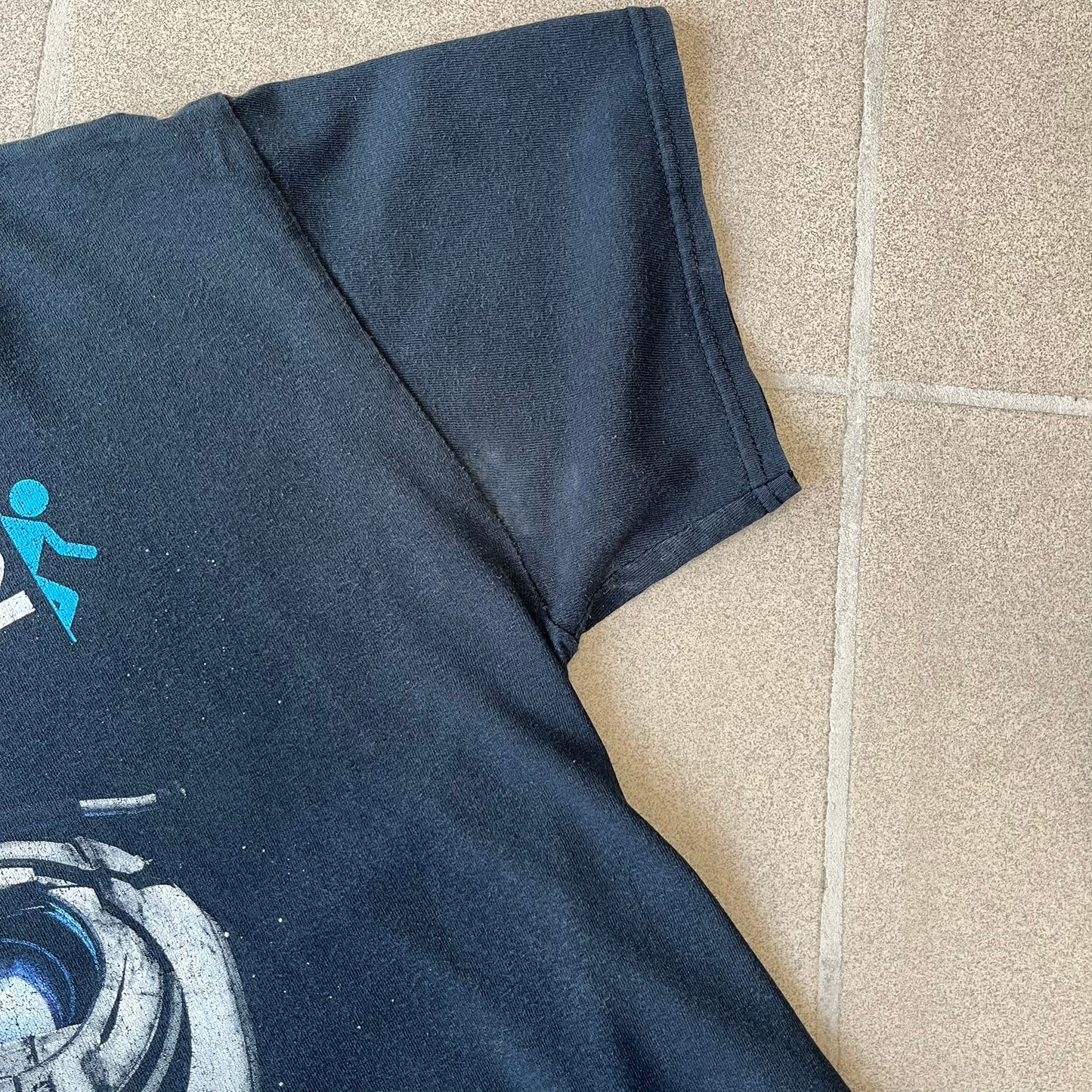 Portal 2: Wheatley in Space 2011 J!nx Tee features a black T-shirt made on a Gildan blank by J!NX in collaboration with Valve. The front has a graphic of Wheatley floating in space with a white and blue Portal 2 logo above him. Made from 100% cotton.
