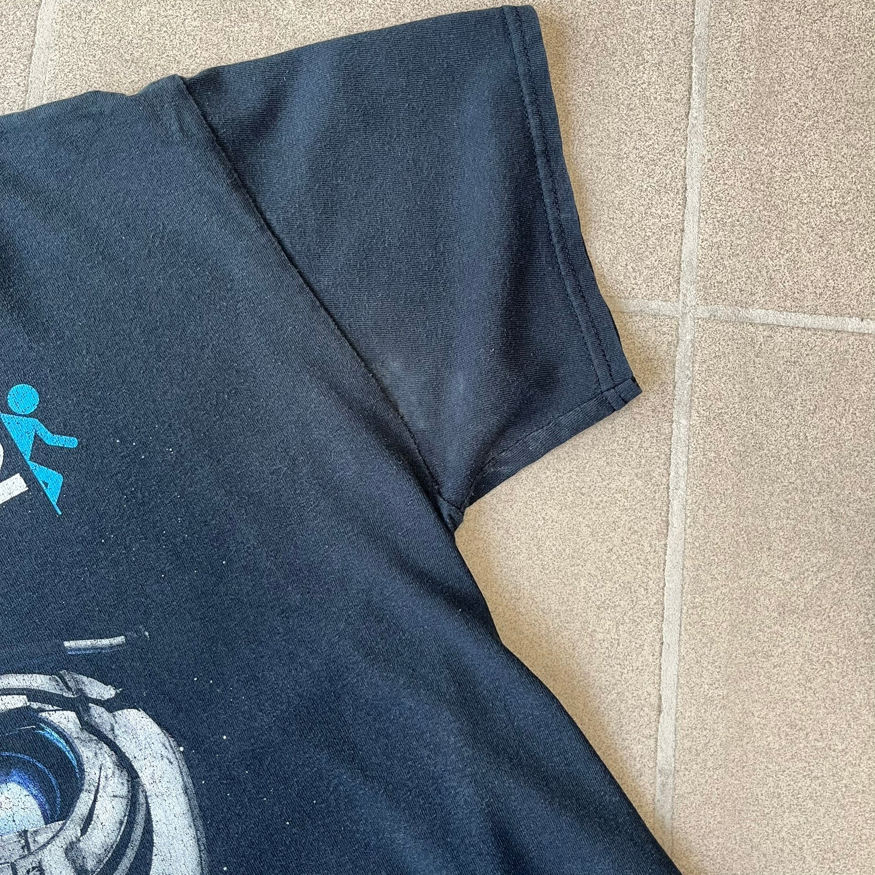 Portal 2: Wheatley in Space 2011 J!nx Tee features a black T-shirt made on a Gildan blank by J!NX in collaboration with Valve. The front has a graphic of Wheatley floating in space with a white and blue Portal 2 logo above him. Made from 100% cotton.
