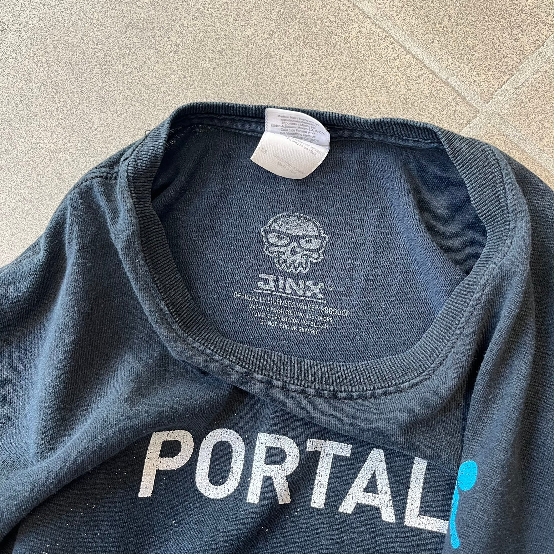 Portal 2: Wheatley in Space 2011 J!nx Tee features a black T-shirt made on a Gildan blank by J!NX in collaboration with Valve. The front has a graphic of Wheatley floating in space with a white and blue Portal 2 logo above him. Made from 100% cotton.
