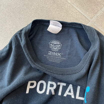 Portal 2: Wheatley in Space 2011 J!nx Tee features a black T-shirt made on a Gildan blank by J!NX in collaboration with Valve. The front has a graphic of Wheatley floating in space with a white and blue Portal 2 logo above him. Made from 100% cotton.

