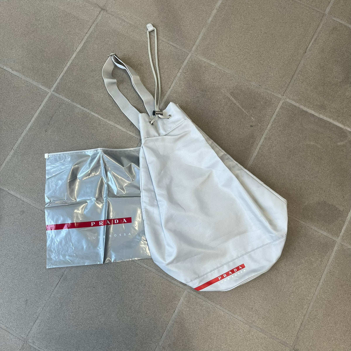 Prada 2012 Luna Rossa Drawstring Sling Bag is a light gray, almost silvery sling bag made as a gift-with-purchase for the Luna Rossa fragrance. It features a red rubber stripe at the bottom with white "Prada" and "Luna Rossa" text, and Prada branding on the drawstring toggle.