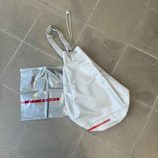 Prada 2012 Luna Rossa Drawstring Sling Bag is a light gray, almost silvery sling bag made as a gift-with-purchase for the Luna Rossa fragrance. It features a red rubber stripe at the bottom with white "Prada" and "Luna Rossa" text, and Prada branding on the drawstring toggle.