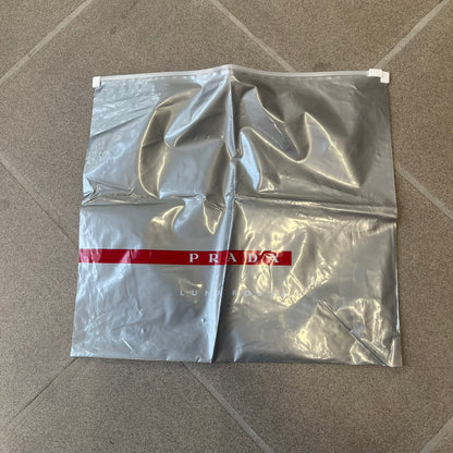 Prada 2012 Luna Rossa Drawstring Sling Bag is a light gray, almost silvery sling bag made as a gift-with-purchase for the Luna Rossa fragrance. It features a red rubber stripe at the bottom with white "Prada" and "Luna Rossa" text, and Prada branding on the drawstring toggle.