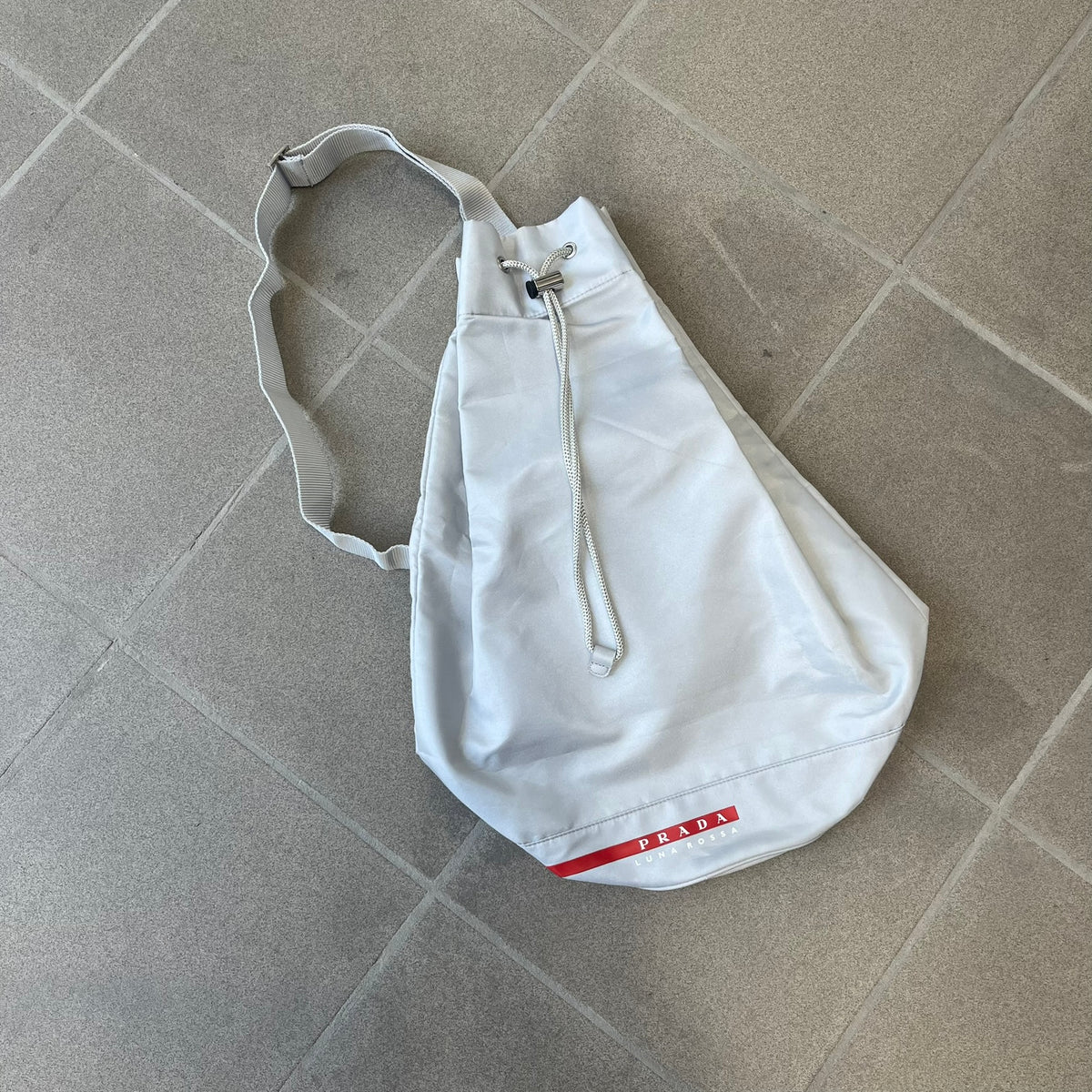 Prada 2012 Luna Rossa Drawstring Sling Bag is a light gray, almost silvery sling bag made as a gift-with-purchase for the Luna Rossa fragrance. It features a red rubber stripe at the bottom with white "Prada" and "Luna Rossa" text, and Prada branding on the drawstring toggle.