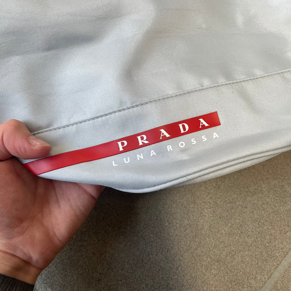 Prada 2012 Luna Rossa Drawstring Sling Bag is a light gray, almost silvery sling bag made as a gift-with-purchase for the Luna Rossa fragrance. It features a red rubber stripe at the bottom with white "Prada" and "Luna Rossa" text, and Prada branding on the drawstring toggle.