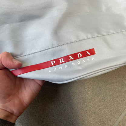 Prada 2012 Luna Rossa Drawstring Sling Bag is a light gray, almost silvery sling bag made as a gift-with-purchase for the Luna Rossa fragrance. It features a red rubber stripe at the bottom with white "Prada" and "Luna Rossa" text, and Prada branding on the drawstring toggle.
