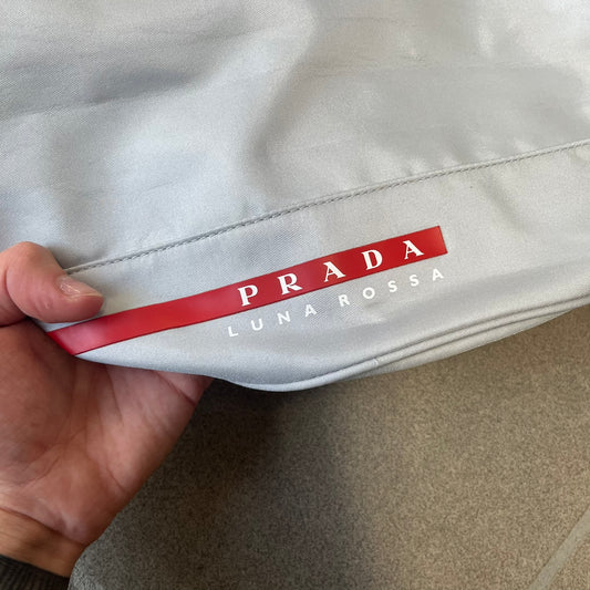 Prada 2012 Luna Rossa Drawstring Sling Bag is a light gray, almost silvery sling bag made as a gift-with-purchase for the Luna Rossa fragrance. It features a red rubber stripe at the bottom with white "Prada" and "Luna Rossa" text, and Prada branding on the drawstring toggle.