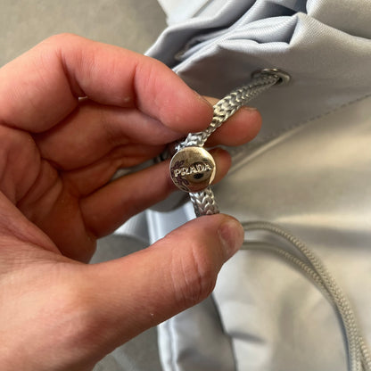 Prada 2012 Luna Rossa Drawstring Sling Bag is a light gray, almost silvery sling bag made as a gift-with-purchase for the Luna Rossa fragrance. It features a red rubber stripe at the bottom with white "Prada" and "Luna Rossa" text, and Prada branding on the drawstring toggle.