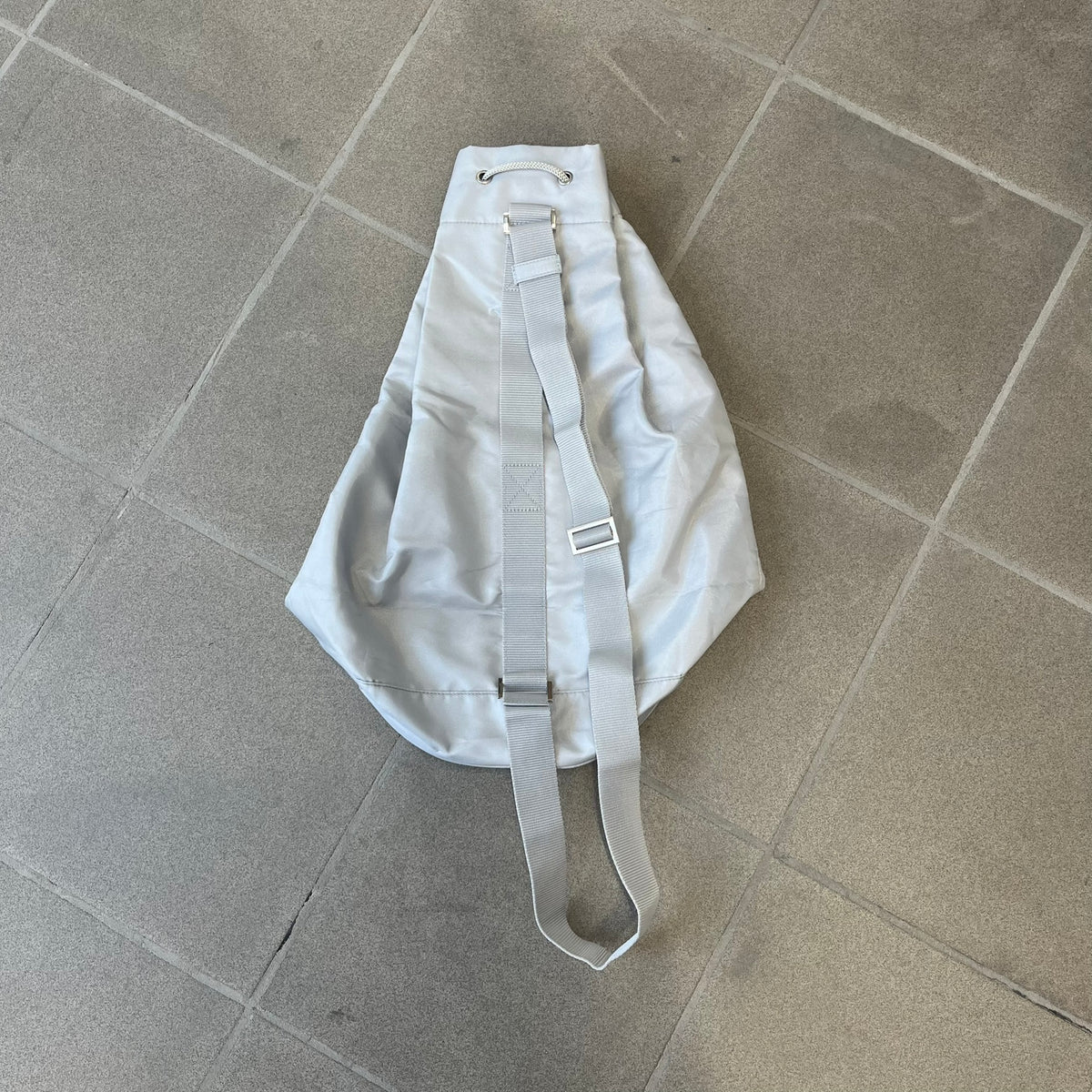 Prada 2012 Luna Rossa Drawstring Sling Bag is a light gray, almost silvery sling bag made as a gift-with-purchase for the Luna Rossa fragrance. It features a red rubber stripe at the bottom with white "Prada" and "Luna Rossa" text, and Prada branding on the drawstring toggle.