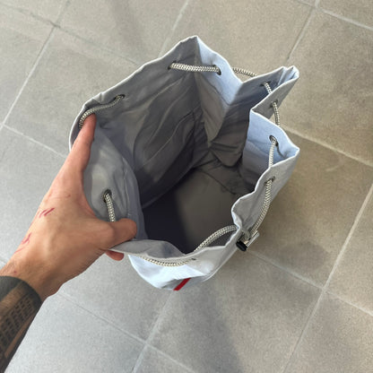 Prada 2012 Luna Rossa Drawstring Sling Bag is a light gray, almost silvery sling bag made as a gift-with-purchase for the Luna Rossa fragrance. It features a red rubber stripe at the bottom with white "Prada" and "Luna Rossa" text, and Prada branding on the drawstring toggle.