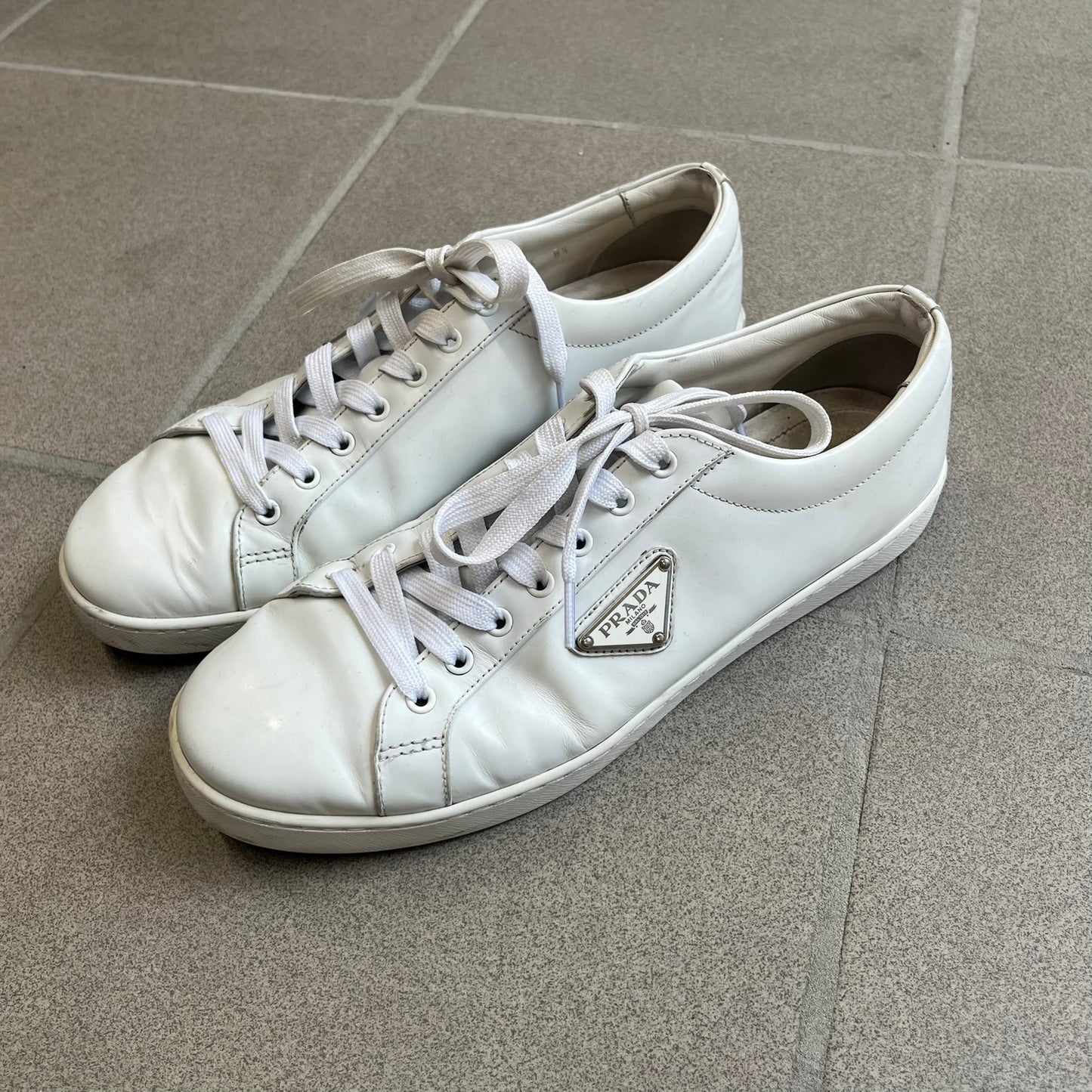 Prada Brushed Leather 2022 White Sneakers feature a classic low-top silhouette crafted from smooth white brushed leather. The outer sides are detailed with Prada's iconic enamel triangle logo plaques in white and silver-tone metal. Made in Italy.