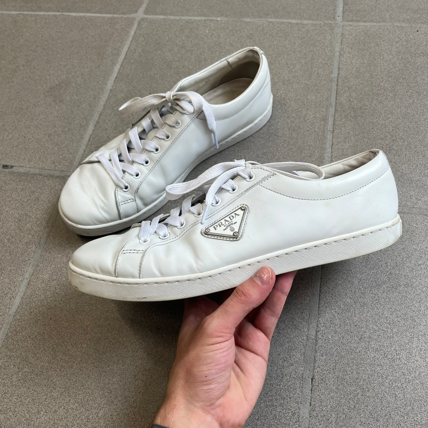 Prada Brushed Leather 2022 White Sneakers feature a classic low-top silhouette crafted from smooth white brushed leather. The outer sides are detailed with Prada's iconic enamel triangle logo plaques in white and silver-tone metal. Made in Italy.