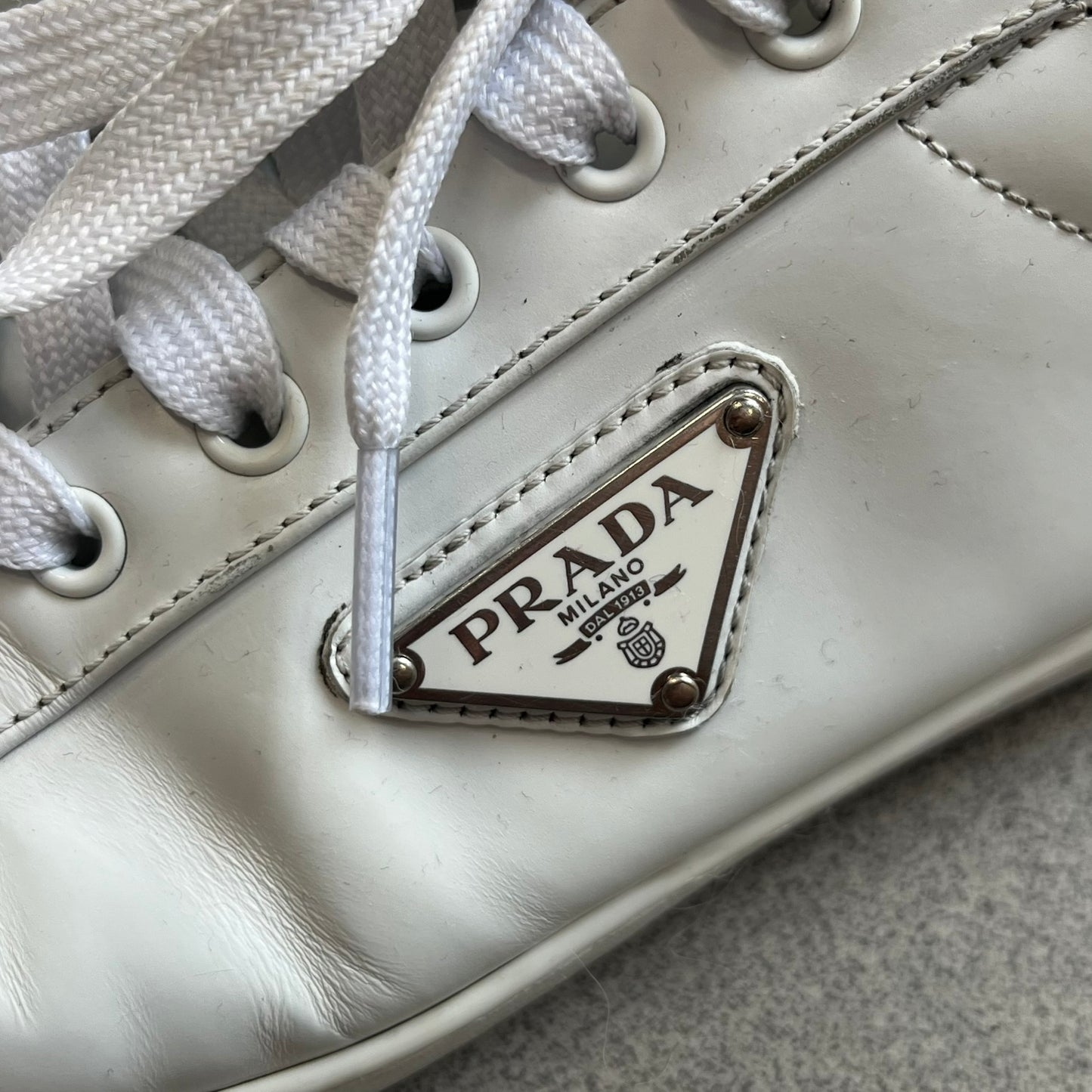 Prada Brushed Leather 2022 White Sneakers feature a classic low-top silhouette crafted from smooth white brushed leather. The outer sides are detailed with Prada's iconic enamel triangle logo plaques in white and silver-tone metal. Made in Italy.