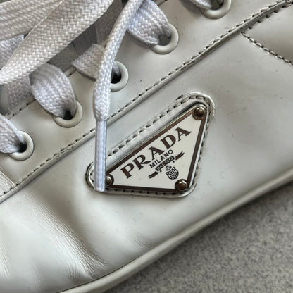 Prada Brushed Leather 2022 White Sneakers feature a classic low-top silhouette crafted from smooth white brushed leather. The outer sides are detailed with Prada's iconic enamel triangle logo plaques in white and silver-tone metal. Made in Italy.