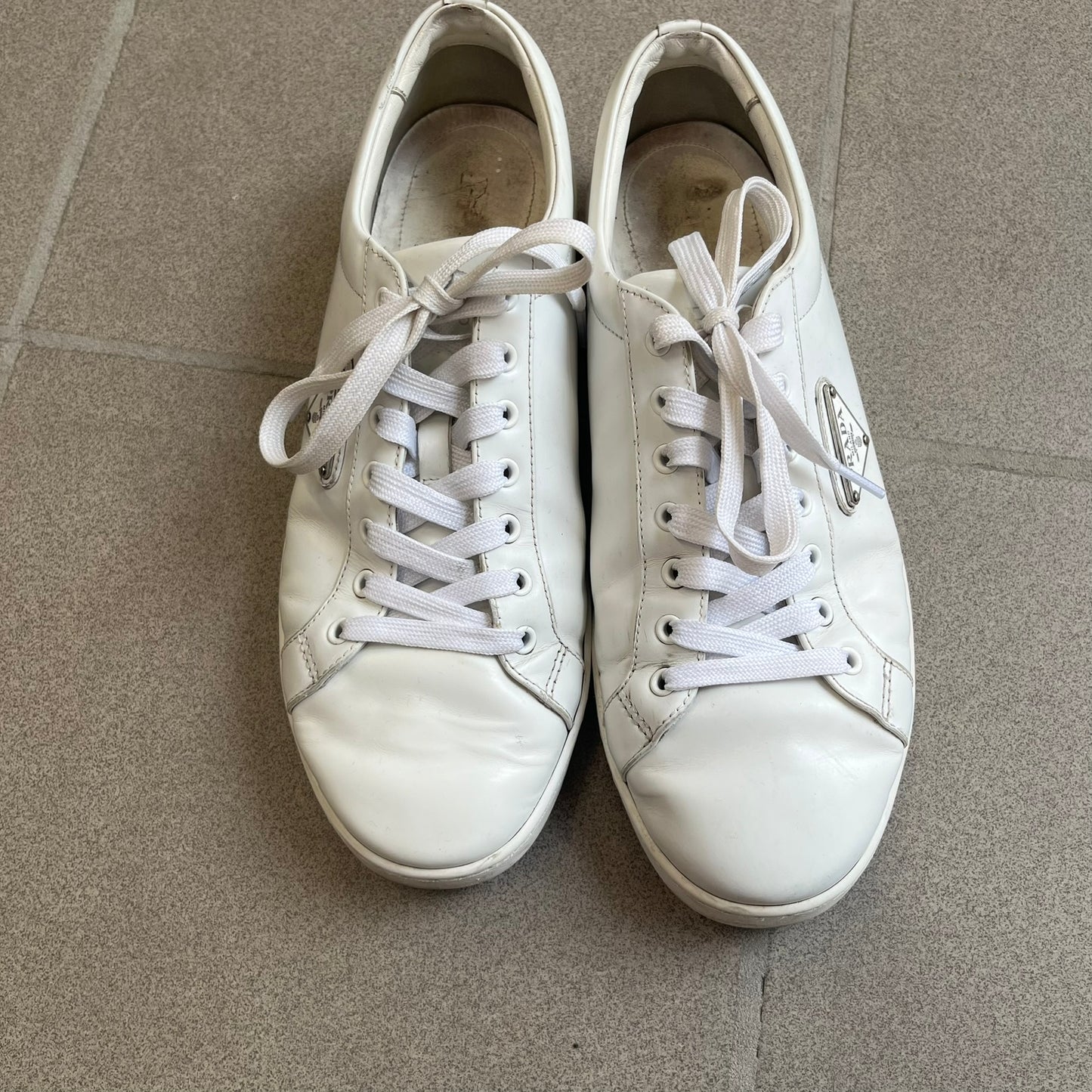 Prada Brushed Leather 2022 White Sneakers feature a classic low-top silhouette crafted from smooth white brushed leather. The outer sides are detailed with Prada's iconic enamel triangle logo plaques in white and silver-tone metal. Made in Italy.