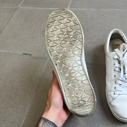 Prada Brushed Leather 2022 White Sneakers feature a classic low-top silhouette crafted from smooth white brushed leather. The outer sides are detailed with Prada's iconic enamel triangle logo plaques in white and silver-tone metal. Made in Italy.