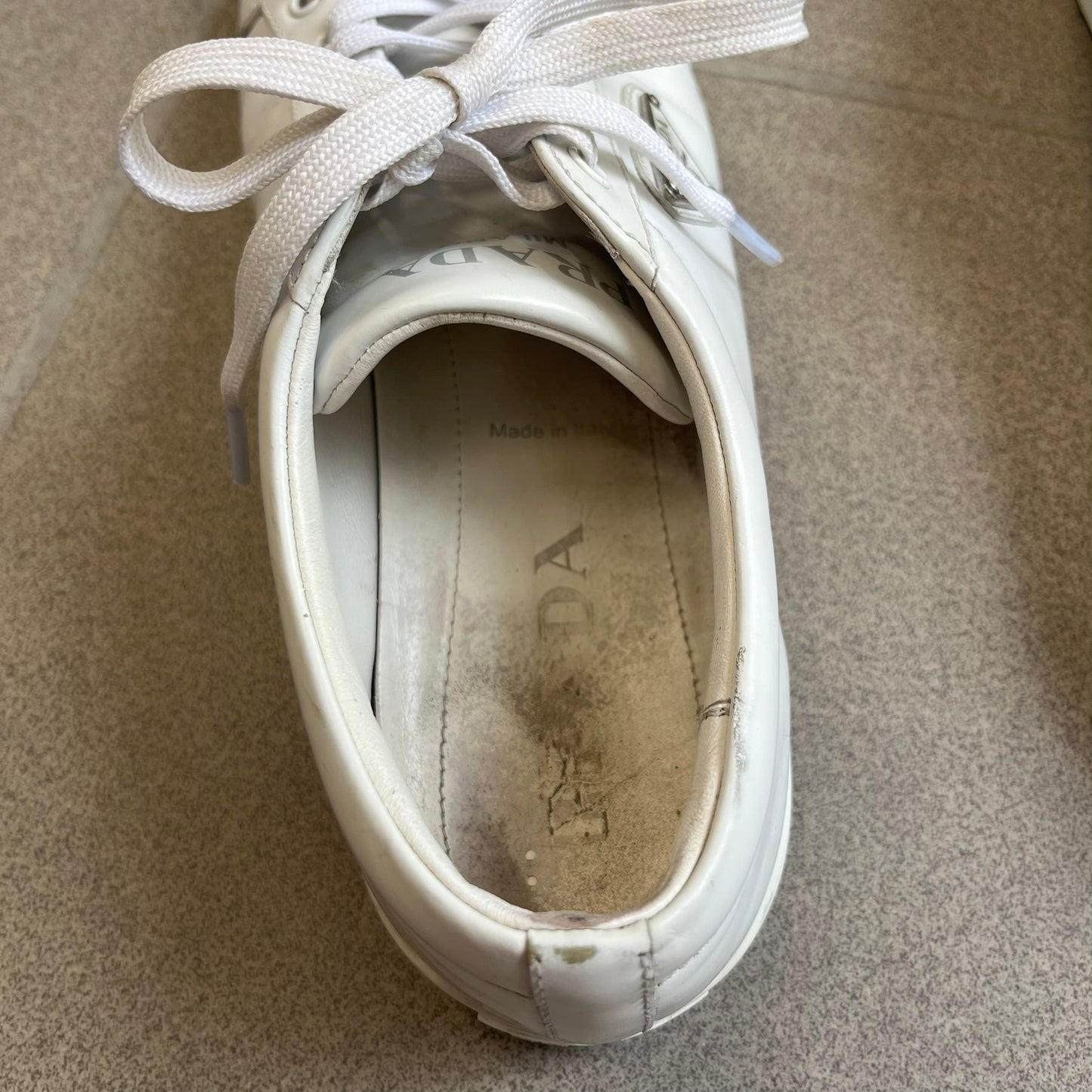 Prada Brushed Leather 2022 White Sneakers feature a classic low-top silhouette crafted from smooth white brushed leather. The outer sides are detailed with Prada's iconic enamel triangle logo plaques in white and silver-tone metal. Made in Italy.