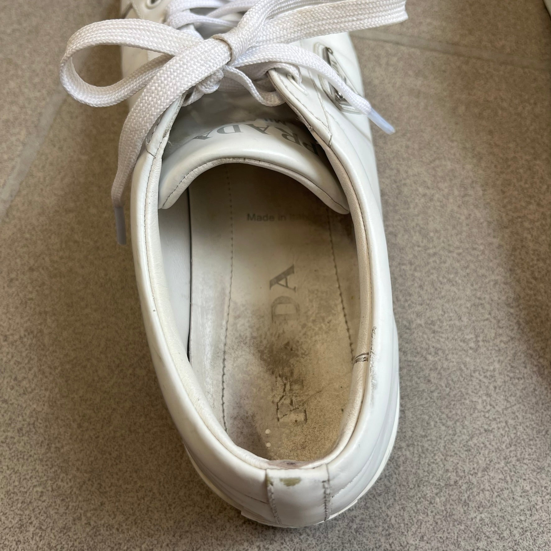 Prada Brushed Leather 2022 White Sneakers feature a classic low-top silhouette crafted from smooth white brushed leather. The outer sides are detailed with Prada's iconic enamel triangle logo plaques in white and silver-tone metal. Made in Italy.