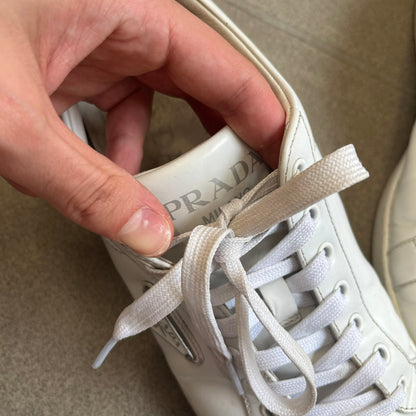 Prada Brushed Leather 2022 White Sneakers feature a classic low-top silhouette crafted from smooth white brushed leather. The outer sides are detailed with Prada's iconic enamel triangle logo plaques in white and silver-tone metal. Made in Italy.