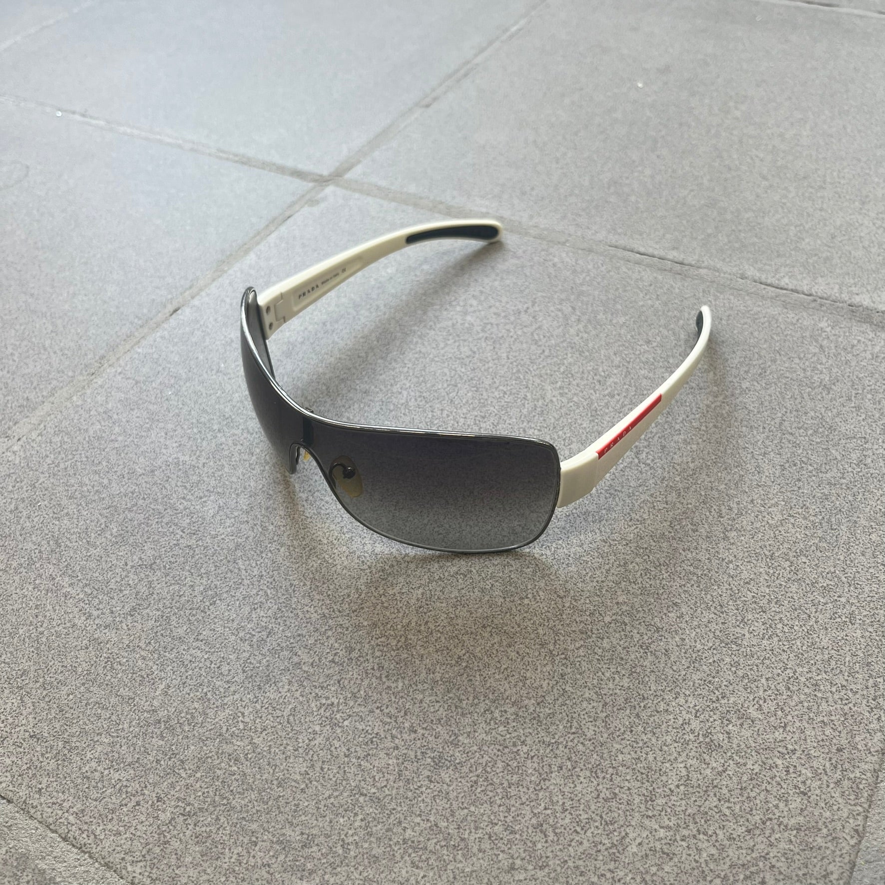 Prada Linea Rossa SPS57G White Sunglasses are shield-style sunglasses featuring a single dark wraparound lens and white arms. One temple is finished with the signature red Prada Linea Rossa logo insert, while the nose pads feature subtle Prada branding. Made in Italy, the sunglasses are lightweight sporty design&nbsp;and they come complete with the original Prada case.