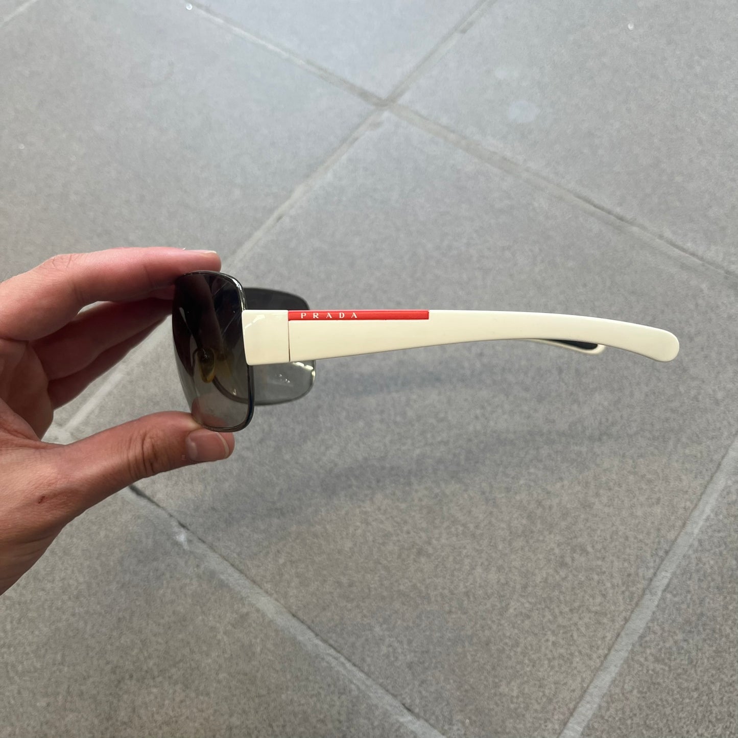 Prada Linea Rossa SPS57G White Sunglasses are shield-style sunglasses featuring a single dark wraparound lens and white arms. One temple is finished with the signature red Prada Linea Rossa logo insert, while the nose pads feature subtle Prada branding. Made in Italy, the sunglasses are lightweight sporty design&nbsp;and they come complete with the original Prada case.