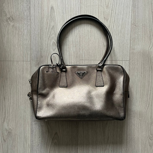 Vintage Prada Metallic Saffiano Bauletto Bag crafted from metallic gold saffiano leather embodies vintage elegance and modern luxury. Detailed with gold-tone hardware, dual handles, and original accessories, it’s a testament to Prada’s enduring craftsmanship. A collector’s piece made to shine on any occasion.