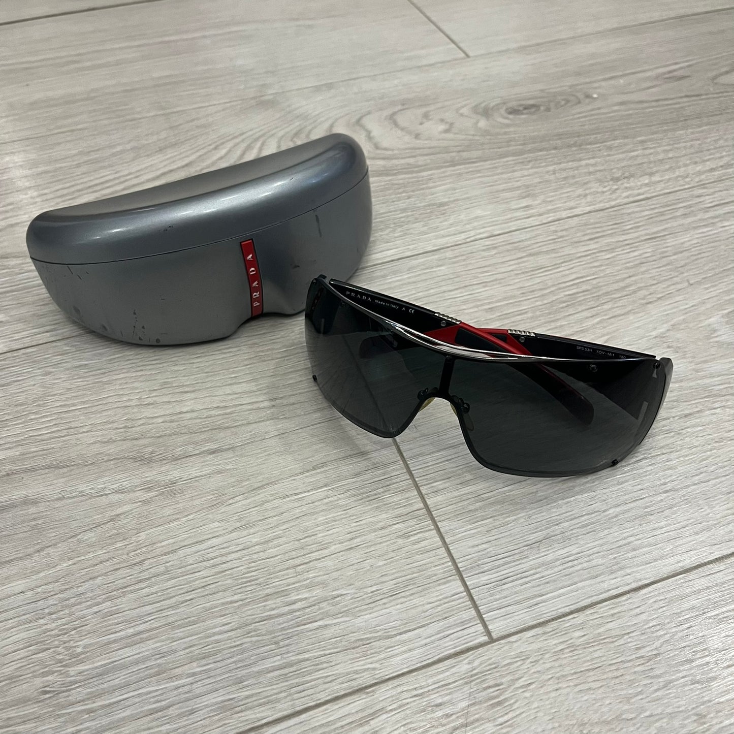 Prada SPS 53H Black/Silver Sunglasses are a vintage wraparound pair featuring a black frame with gunmetal silver accents. The gray lenses are complemented by red details on the arms and visible screws on the front, with Prada branding on the temples. Crafted in Italy from metal and resin, they come complete with the original Prada case.