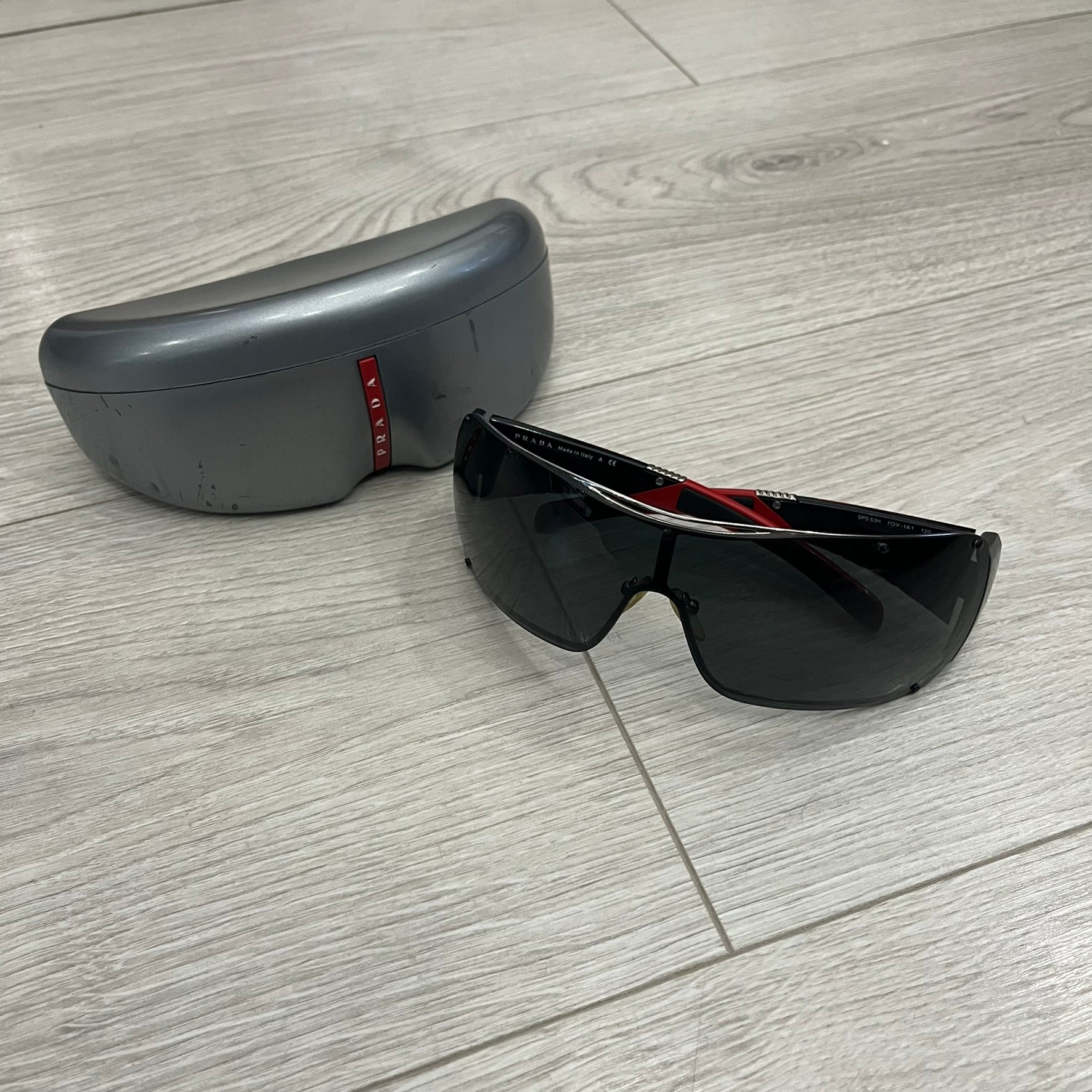 Prada SPS 53H Black/Silver Sunglasses are a vintage wraparound pair featuring a black frame with gunmetal silver accents. The gray lenses are complemented by red details on the arms and visible screws on the front, with Prada branding on the temples. Crafted in Italy from metal and resin, they come complete with the original Prada case.