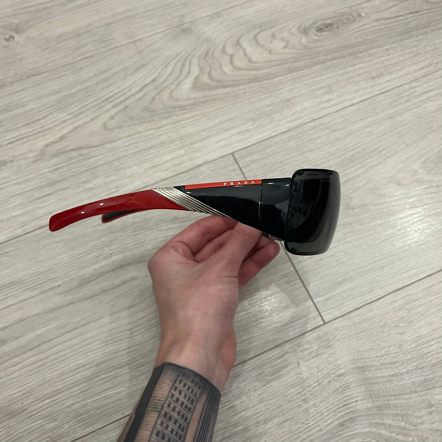 Prada SPS 53H Black/Silver Sunglasses are a vintage wraparound pair featuring a black frame with gunmetal silver accents. The gray lenses are complemented by red details on the arms and visible screws on the front, with Prada branding on the temples. Crafted in Italy from metal and resin, they come complete with the original Prada case.