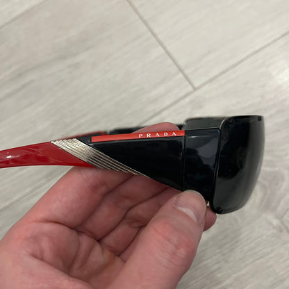 Prada SPS 53H Black/Silver Sunglasses are a vintage wraparound pair featuring a black frame with gunmetal silver accents. The gray lenses are complemented by red details on the arms and visible screws on the front, with Prada branding on the temples. Crafted in Italy from metal and resin, they come complete with the original Prada case.