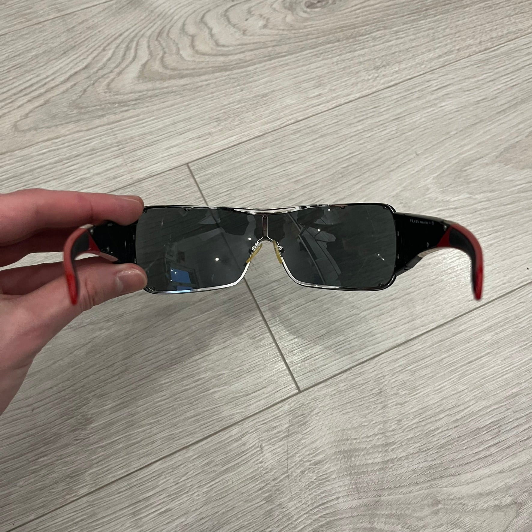 Prada SPS 53H Black/Silver Sunglasses are a vintage wraparound pair featuring a black frame with gunmetal silver accents. The gray lenses are complemented by red details on the arms and visible screws on the front, with Prada branding on the temples. Crafted in Italy from metal and resin, they come complete with the original Prada case.