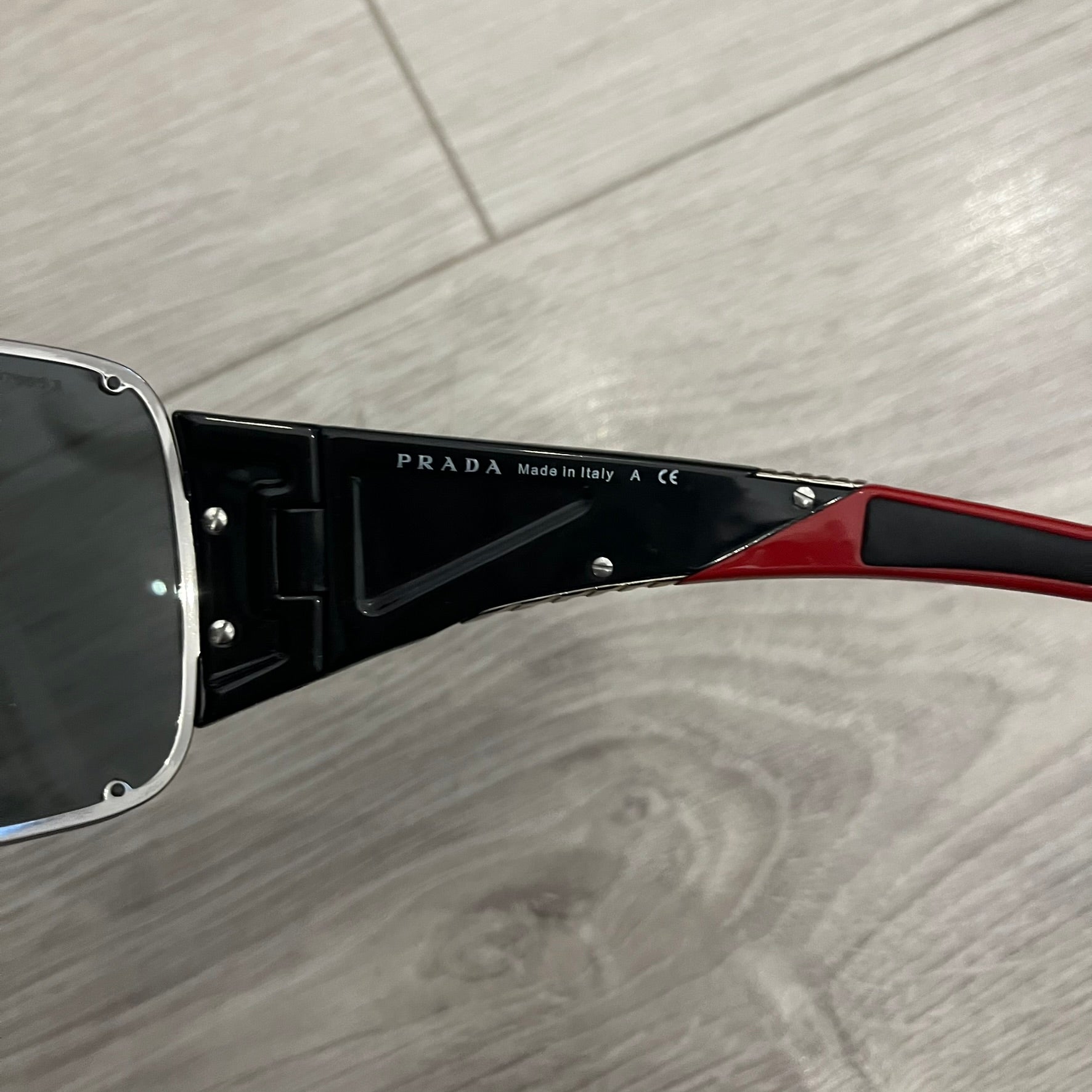 Prada SPS 53H Black/Silver Sunglasses are a vintage wraparound pair featuring a black frame with gunmetal silver accents. The gray lenses are complemented by red details on the arms and visible screws on the front, with Prada branding on the temples. Crafted in Italy from metal and resin, they come complete with the original Prada case.
