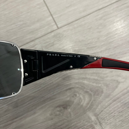 Prada SPS 53H Black/Silver Sunglasses are a vintage wraparound pair featuring a black frame with gunmetal silver accents. The gray lenses are complemented by red details on the arms and visible screws on the front, with Prada branding on the temples. Crafted in Italy from metal and resin, they come complete with the original Prada case.