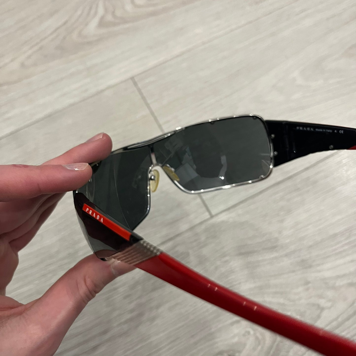 Prada SPS 53H Black/Silver Sunglasses are a vintage wraparound pair featuring a black frame with gunmetal silver accents. The gray lenses are complemented by red details on the arms and visible screws on the front, with Prada branding on the temples. Crafted in Italy from metal and resin, they come complete with the original Prada case.