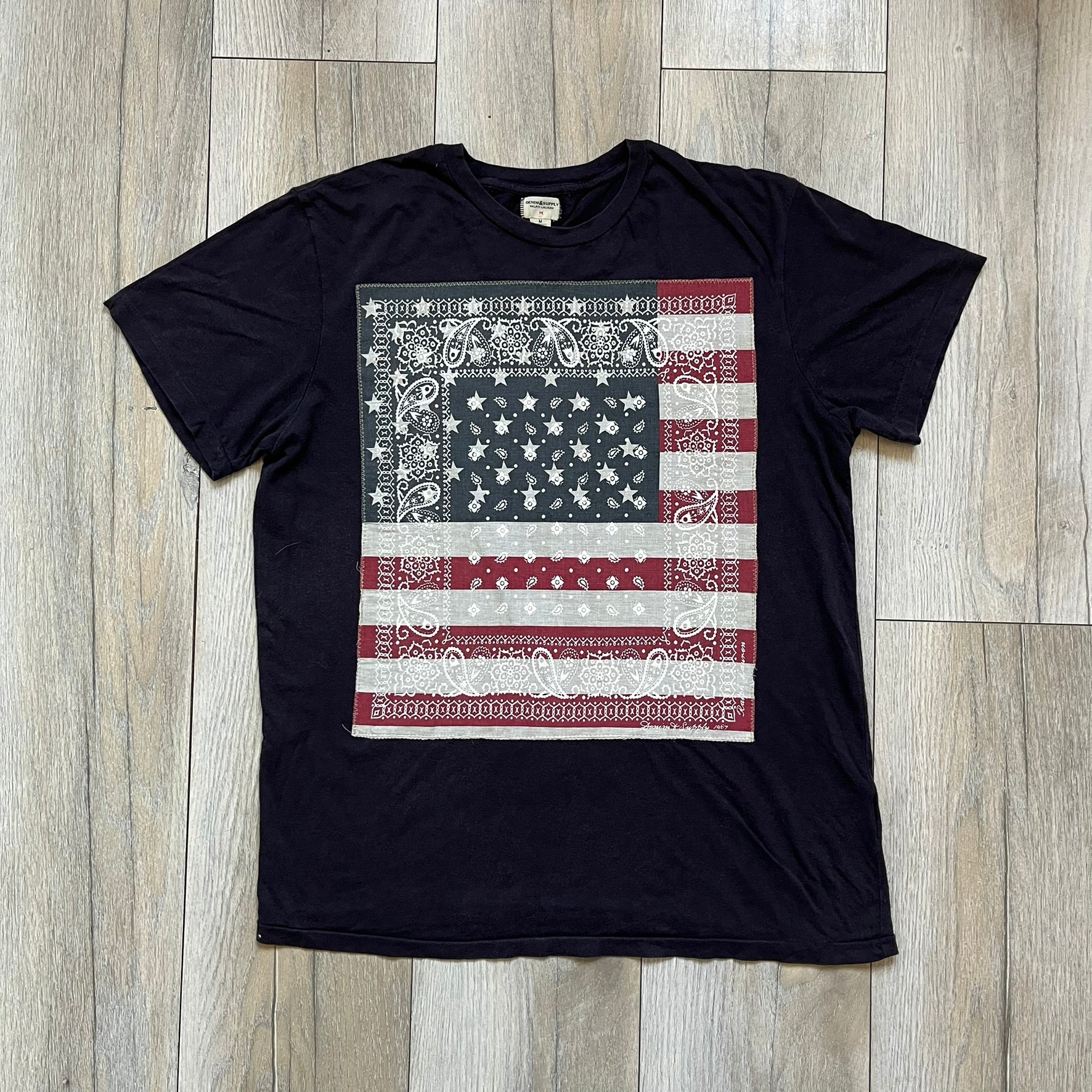 Ralph Lauren 2000s USA Bandana Flag Tee is a navy short-sleeve T-shirt made from 100% cotton. The front features a large USA flag constructed from fabric with a white bandana-style pattern, sewn onto the shirt for a raised effect. This piece comes from the Ralph Lauren Denim &amp; Supply line.