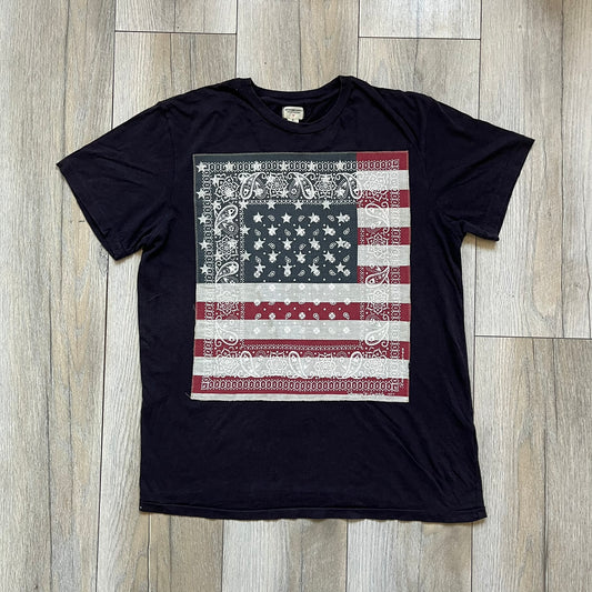 Ralph Lauren 2000s USA Bandana Flag Tee is a navy short-sleeve T-shirt made from 100% cotton. The front features a large USA flag constructed from fabric with a white bandana-style pattern, sewn onto the shirt for a raised effect. This piece comes from the Ralph Lauren Denim &amp; Supply line.