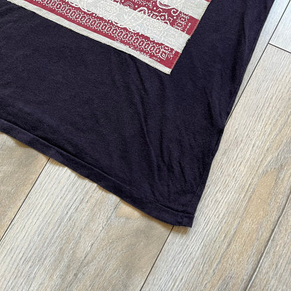 Ralph Lauren 2000s USA Bandana Flag Tee is a navy short-sleeve T-shirt made from 100% cotton. The front features a large USA flag constructed from fabric with a white bandana-style pattern, sewn onto the shirt for a raised effect. This piece comes from the Ralph Lauren Denim &amp; Supply line.