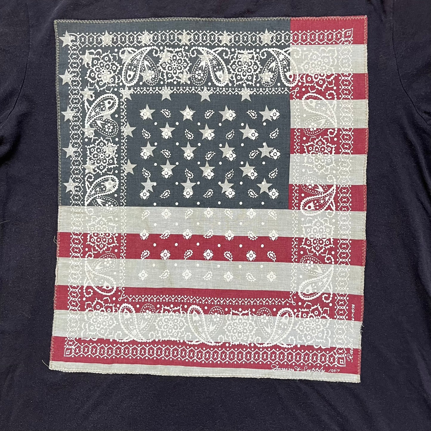 Ralph Lauren 2000s USA Bandana Flag Tee is a navy short-sleeve T-shirt made from 100% cotton. The front features a large USA flag constructed from fabric with a white bandana-style pattern, sewn onto the shirt for a raised effect. This piece comes from the Ralph Lauren Denim &amp; Supply line.