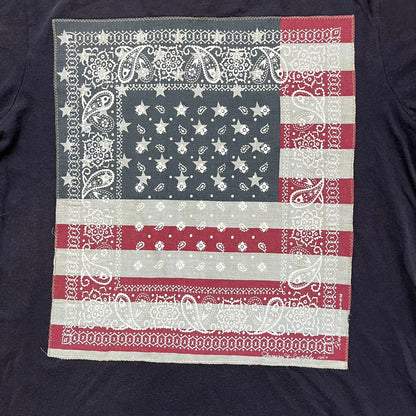 Ralph Lauren 2000s USA Bandana Flag Tee is a navy short-sleeve T-shirt made from 100% cotton. The front features a large USA flag constructed from fabric with a white bandana-style pattern, sewn onto the shirt for a raised effect. This piece comes from the Ralph Lauren Denim &amp; Supply line.