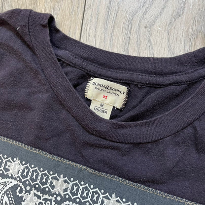 Ralph Lauren 2000s USA Bandana Flag Tee is a navy short-sleeve T-shirt made from 100% cotton. The front features a large USA flag constructed from fabric with a white bandana-style pattern, sewn onto the shirt for a raised effect. This piece comes from the Ralph Lauren Denim &amp; Supply line.