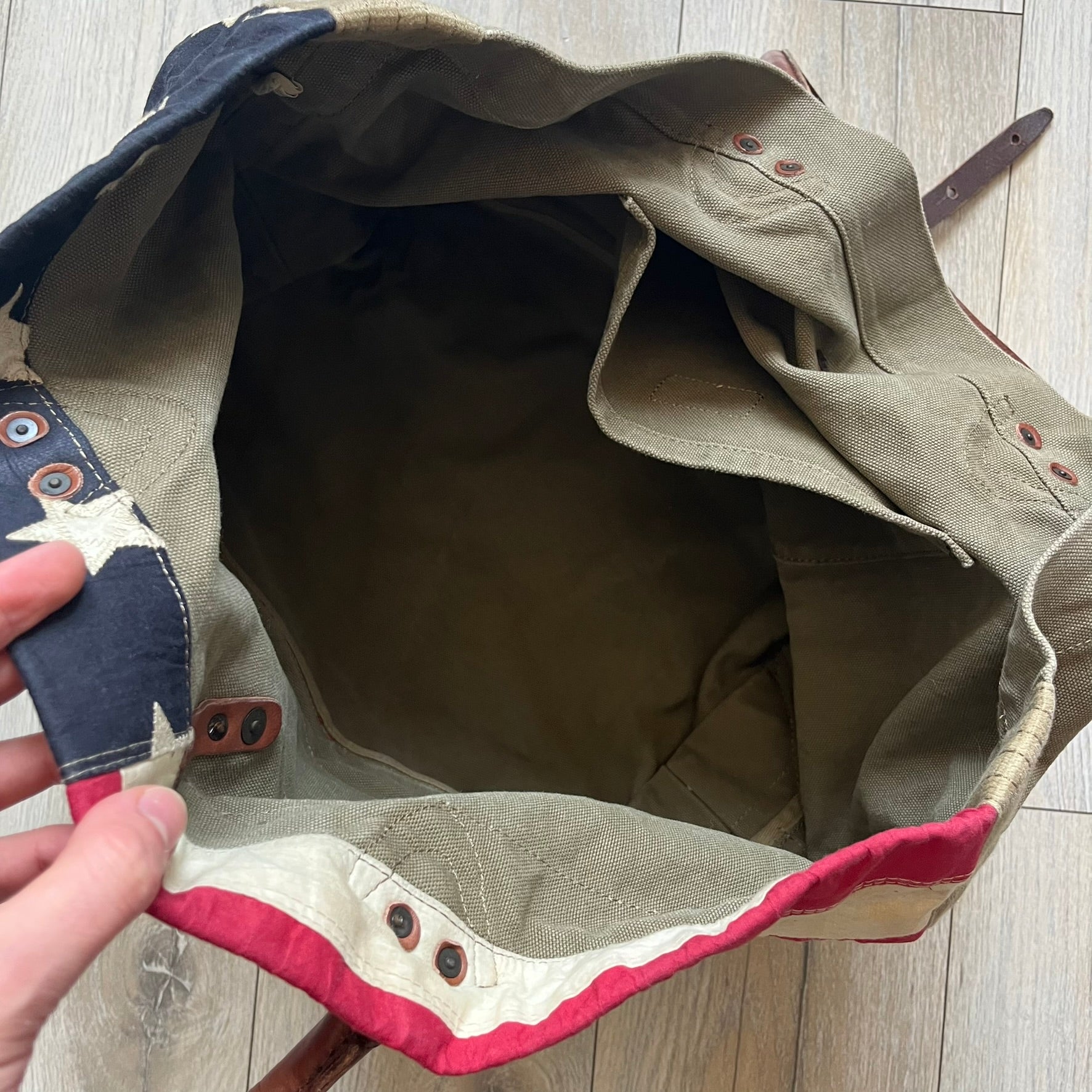 Ralph Lauren Denim &amp; Supply 1990s USA Flag Shoulder Bag is a large shoulder bag constructed from a mix of leather and cotton canvas. One side features a faded USA flag made from stitched canvas patches, while the reverse side is khaki canvas. The bag includes leather handles and a leather strap closure, an interior main compartment with additional smaller pockets, and a Denim &amp; Supply tag.
