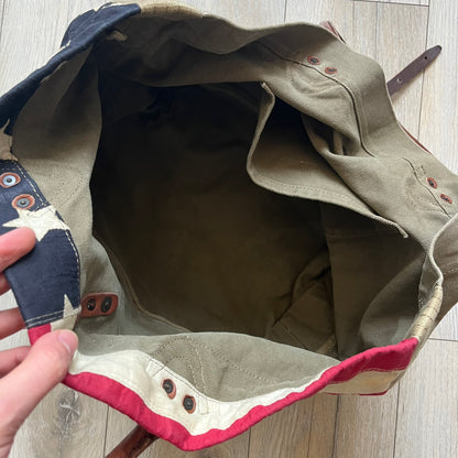 Ralph Lauren Denim &amp; Supply 1990s USA Flag Shoulder Bag is a large shoulder bag constructed from a mix of leather and cotton canvas. One side features a faded USA flag made from stitched canvas patches, while the reverse side is khaki canvas. The bag includes leather handles and a leather strap closure, an interior main compartment with additional smaller pockets, and a Denim &amp; Supply tag.
