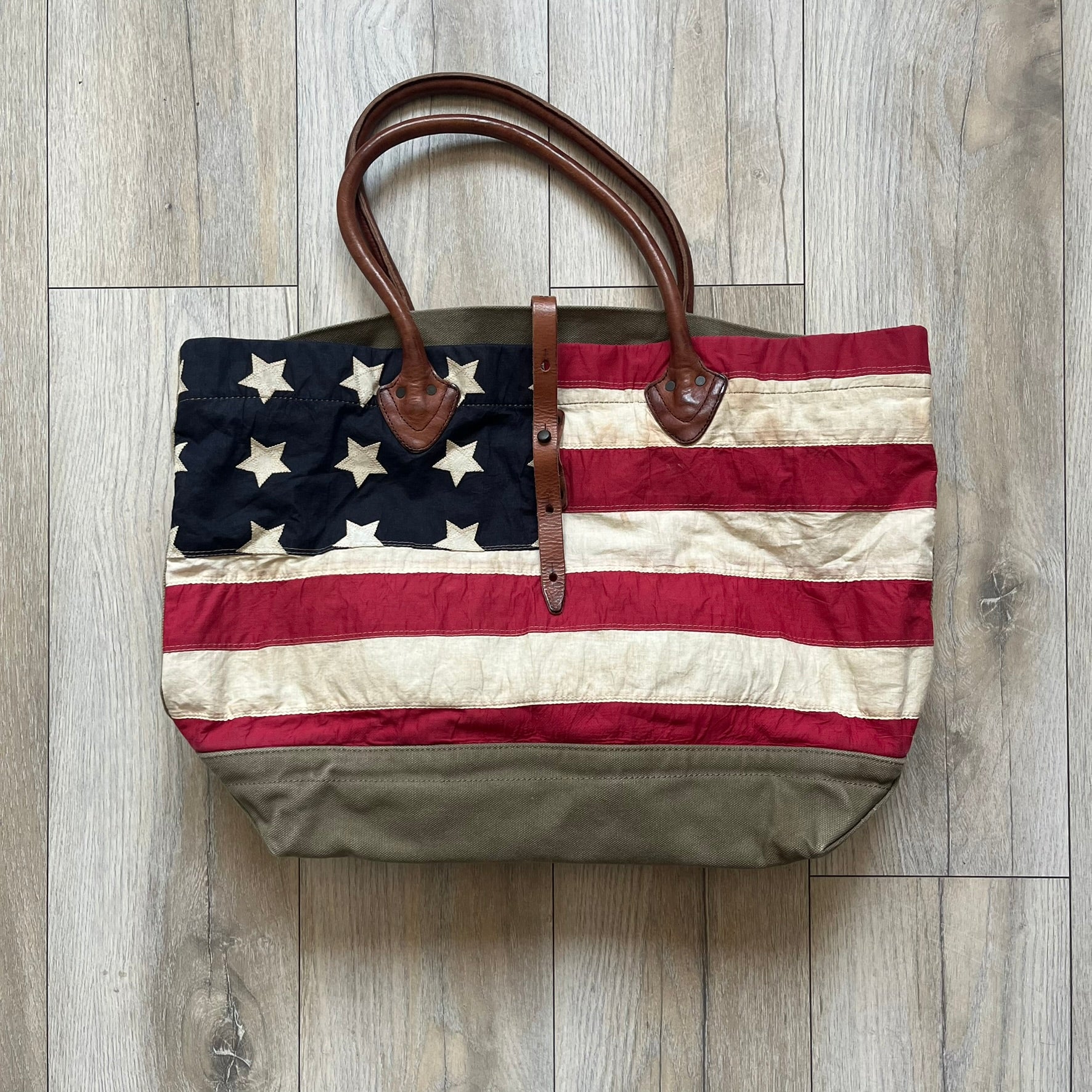 Ralph Lauren Denim &amp; Supply 1990s USA Flag Shoulder Bag is a large shoulder bag constructed from a mix of leather and cotton canvas. One side features a faded USA flag made from stitched canvas patches, while the reverse side is khaki canvas. The bag includes leather handles and a leather strap closure, an interior main compartment with additional smaller pockets, and a Denim &amp; Supply tag.
