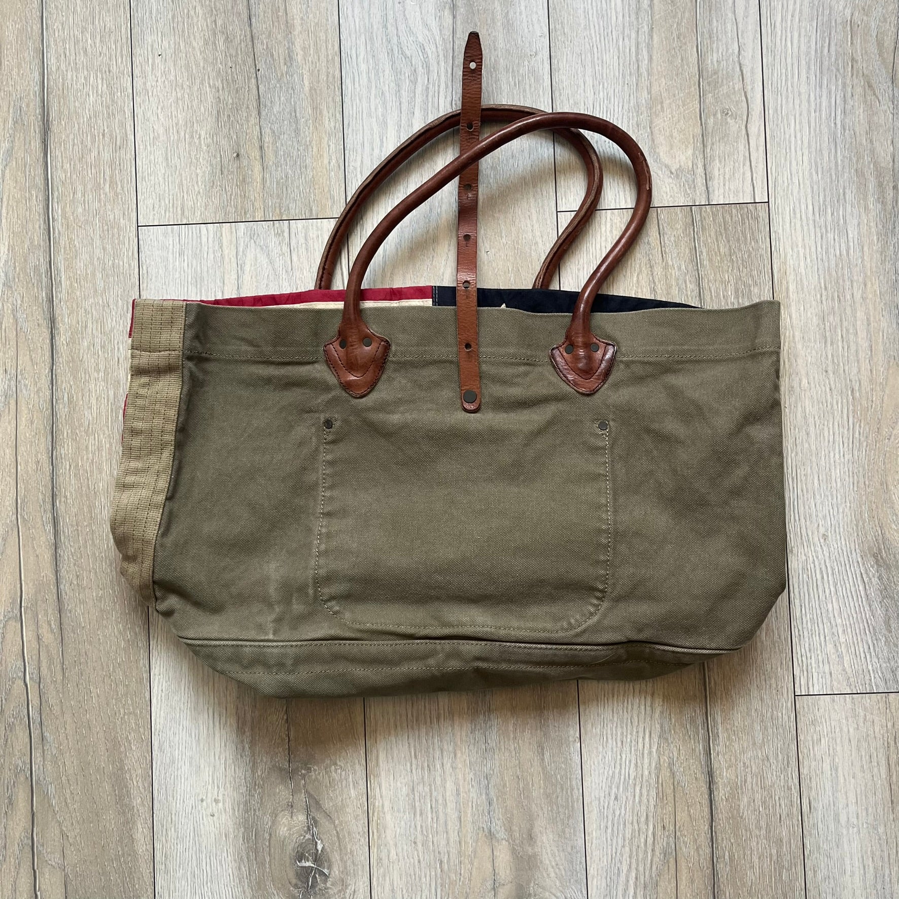 Ralph Lauren Denim &amp; Supply 1990s USA Flag Shoulder Bag is a large shoulder bag constructed from a mix of leather and cotton canvas. One side features a faded USA flag made from stitched canvas patches, while the reverse side is khaki canvas. The bag includes leather handles and a leather strap closure, an interior main compartment with additional smaller pockets, and a Denim &amp; Supply tag.
