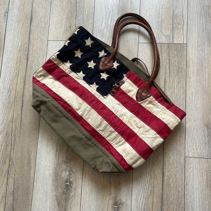 Ralph Lauren Denim &amp; Supply 1990s USA Flag Shoulder Bag is a large shoulder bag constructed from a mix of leather and cotton canvas. One side features a faded USA flag made from stitched canvas patches, while the reverse side is khaki canvas. The bag includes leather handles and a leather strap closure, an interior main compartment with additional smaller pockets, and a Denim &amp; Supply tag.
