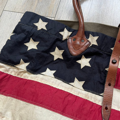 Ralph Lauren Denim &amp; Supply 1990s USA Flag Shoulder Bag is a large shoulder bag constructed from a mix of leather and cotton canvas. One side features a faded USA flag made from stitched canvas patches, while the reverse side is khaki canvas. The bag includes leather handles and a leather strap closure, an interior main compartment with additional smaller pockets, and a Denim &amp; Supply tag.
