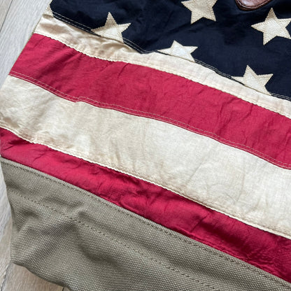 Ralph Lauren Denim &amp; Supply 1990s USA Flag Shoulder Bag is a large shoulder bag constructed from a mix of leather and cotton canvas. One side features a faded USA flag made from stitched canvas patches, while the reverse side is khaki canvas. The bag includes leather handles and a leather strap closure, an interior main compartment with additional smaller pockets, and a Denim &amp; Supply tag.
