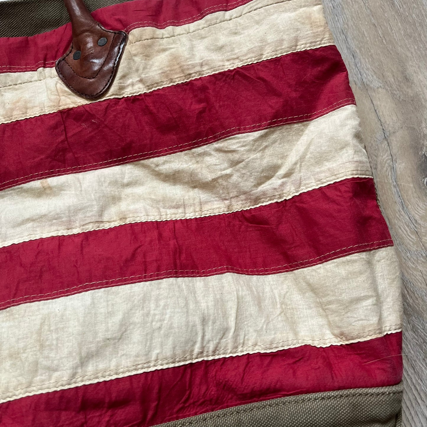 Ralph Lauren Denim &amp; Supply 1990s USA Flag Shoulder Bag is a large shoulder bag constructed from a mix of leather and cotton canvas. One side features a faded USA flag made from stitched canvas patches, while the reverse side is khaki canvas. The bag includes leather handles and a leather strap closure, an interior main compartment with additional smaller pockets, and a Denim &amp; Supply tag.
