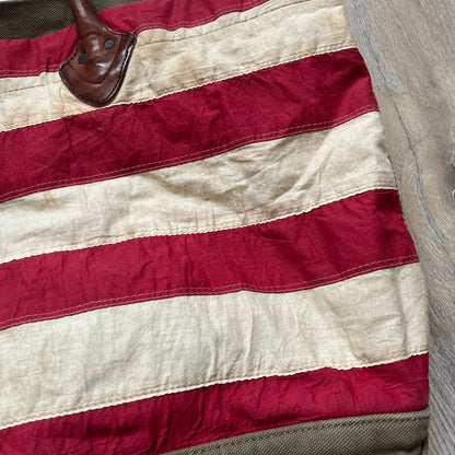 Ralph Lauren Denim &amp; Supply 1990s USA Flag Shoulder Bag is a large shoulder bag constructed from a mix of leather and cotton canvas. One side features a faded USA flag made from stitched canvas patches, while the reverse side is khaki canvas. The bag includes leather handles and a leather strap closure, an interior main compartment with additional smaller pockets, and a Denim &amp; Supply tag.
